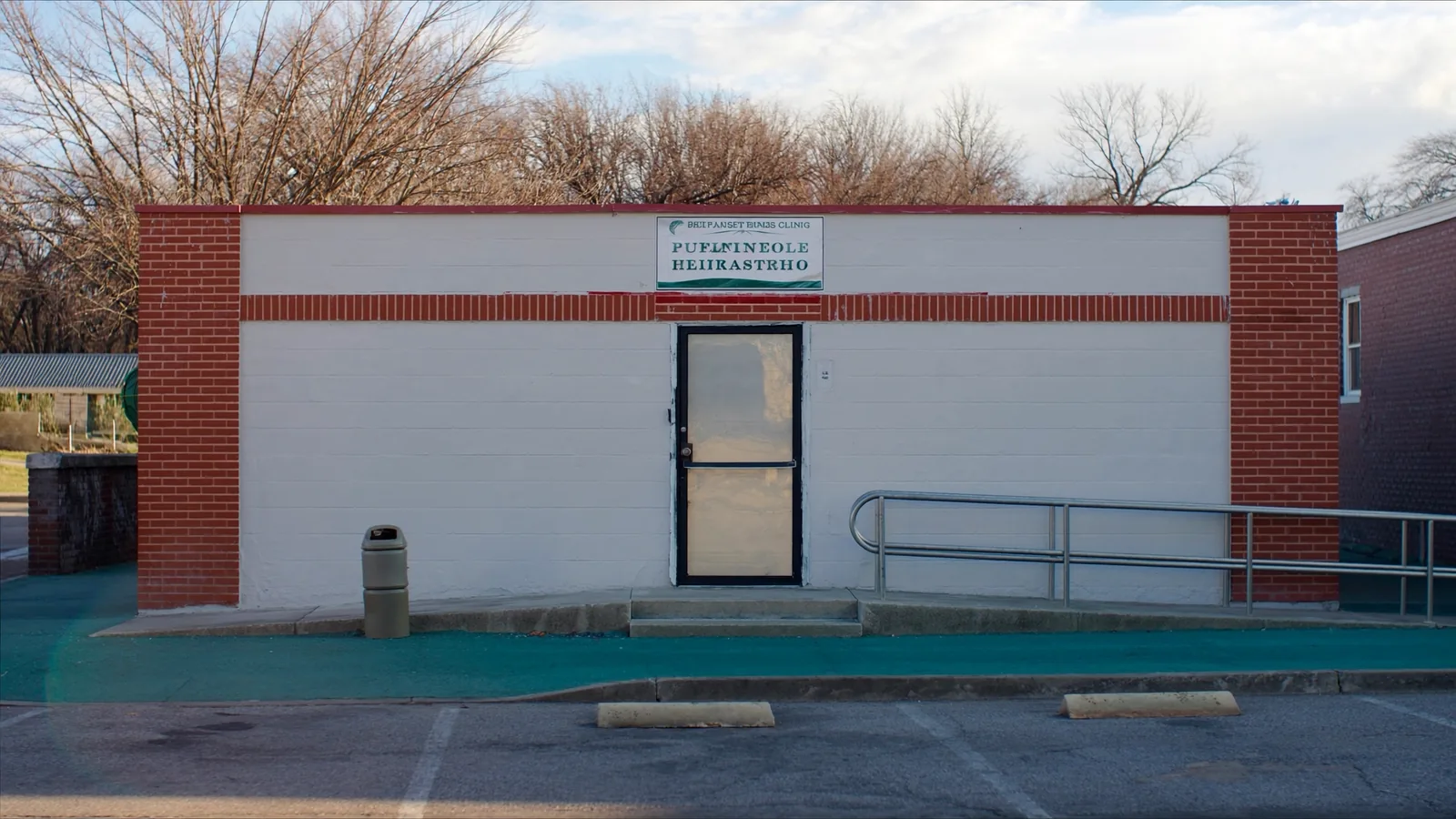 A modest single-story clinic in a converted storefront on a small-town street, with a plain glass door, a wheelchair ramp with a metal handrail and two parking spaces at the kerb.