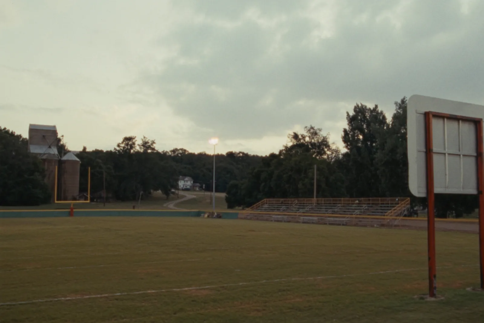 An empty small-town high school football field at dusk with chalked yard lines on worn grass, low aluminium bleachers along one side and floodlights on above a treeline.