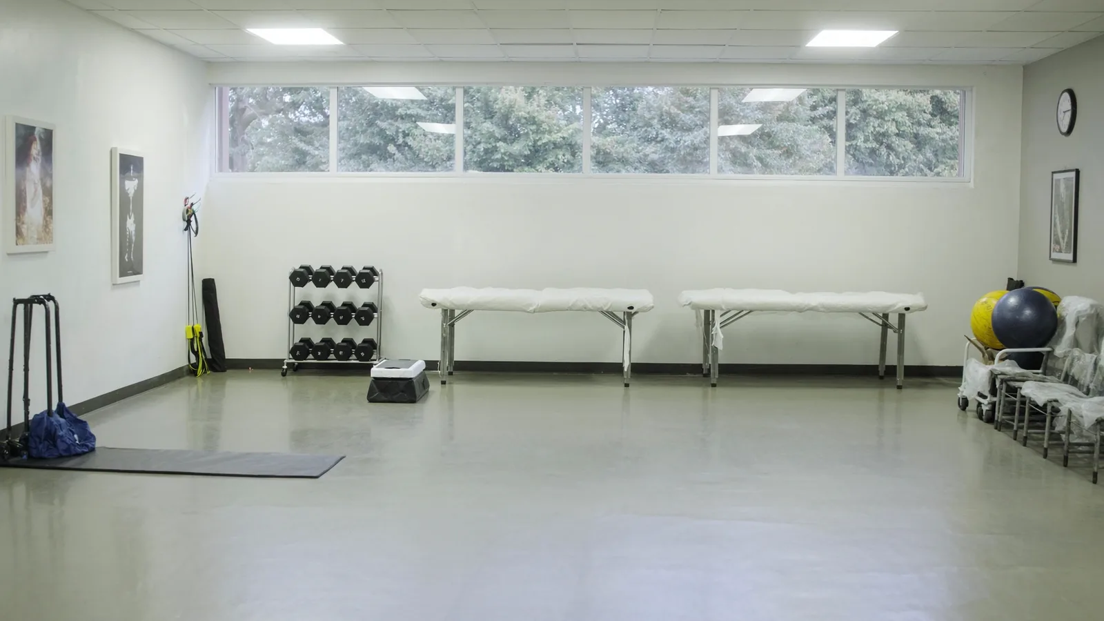 The interior of a small outpatient physical therapy gym with two padded treatment tables, a rack of dumbbells, resistance bands hanging on a hook, a low step box and daylight from windows along one wall.