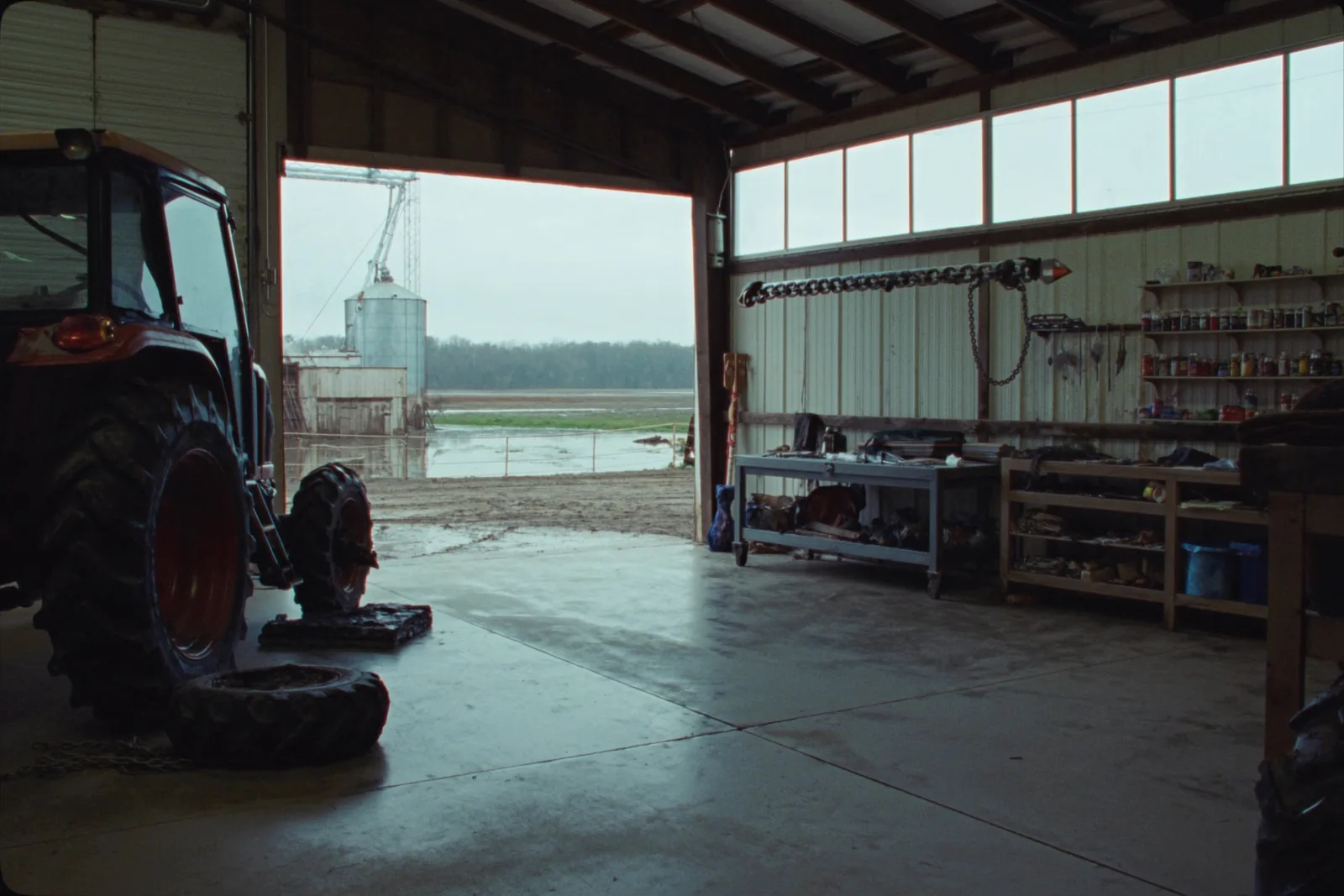 The inside of a working farm shop with a concrete floor, a tractor with one rear wheel removed, a cluttered steel workbench and an open door onto a muddy yard with a grain bin beyond.