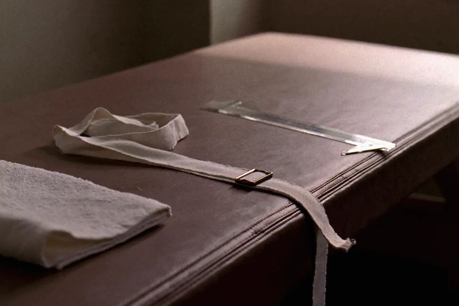 A padded treatment table with a folded towel, a canvas gait belt lying across the edge and a clear plastic goniometer resting on the surface.